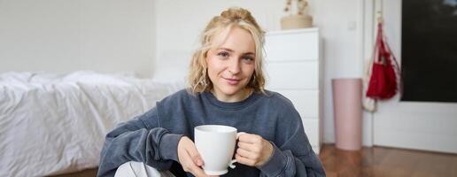 retrato de joven mujer sentado en dormitorio piso, Bebiendo té, participación blanco jarra y sonriente a cámara foto