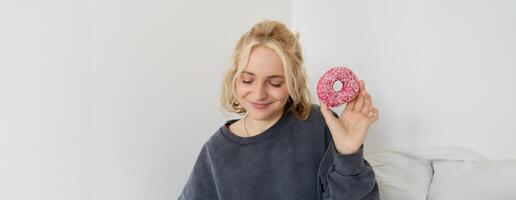 Portrait of cute blond girl holding pink doughnut with sprinkles on top, showing her favourite food photo