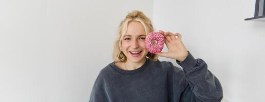 Close up portrait of cute young blond woman, showing pink doughnut near her face, smiling and looking happy photo