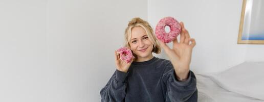 Portrait of beautiful blond woman, showing two pink doughnuts, delicious pastry, eating dessert and smiling, sharing food with you photo