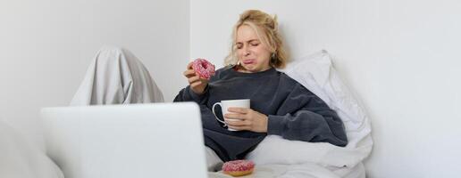 Portrait of woman watching sad movie on laptop, crying and wiping tears off, eating doughnut and drinking tea photo