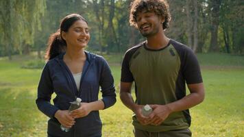 Two sport people smiling talking holding water bottles smile invitation join to exercise male Indian female Arabian friends couple man woman in park invite welcome workout fitness fit in city motivate video