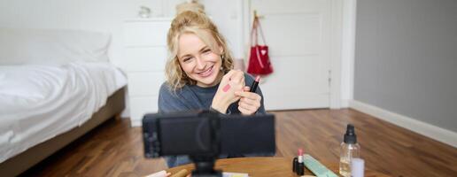 imagen de joven elegante mujer, blogger grabación un belleza estilo de vida de su cosecha mejor lápiz labial, demostración labio bálsamo muestras en su piel, sentado en frente de digital cámara en vacío habitación foto