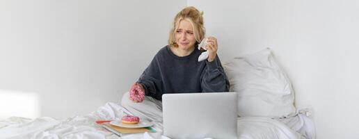 Portrait of woman sitting on a bed with laptop, eating doughnut and crying from sad movie scene photo