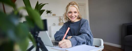 Cute smiling girl sits in a room, writes down notes, doing homework, records of herself on digital camera, creates content for vlog, lifestyle blogger doing daily routine episode photo