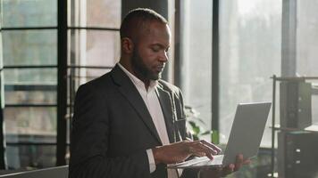 afrikanisch amerikanisch Mann Senior mittleren Alters männlich Geschäftsmann Manager Arbeiten Computer beim Büro sonnig Sonnenstrahl Arbeitsplatz glücklich froh Kerl Geschäft Arbeitgeber Tippen Projekt auf Laptop suchen beim Kamera Lächeln video