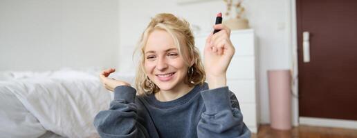 Portrait of young social media influencer, woman recording a with beauty products, showing her makeup on camera, holding lipstick and smiling, sitting on floor photo