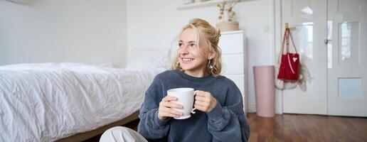 estilo de vida retrato de joven mujer sentado en dormitorio piso con taza de té, Bebiendo desde grande blanco jarra y mirando aparte, sonriente felizmente foto