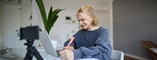 Portrait of smiling blond woman writing in notebook, making notes, recording content for social medial on digital camera, looking at laptop, working or studying photo