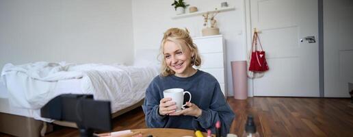 Portrait of beautiful social media beauty blogger, sitting in front of digital camera on floor in bedroom, drinking tea and chatting, talking to followers photo