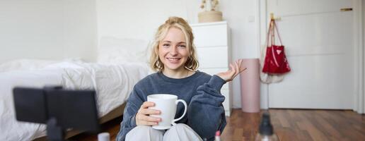 alegre mujer, belleza bloguero, registros estilo de vida vlog en digital cámara, negociaciones por casualidad, dice un historia para social medios de comunicación seguidores, sostiene taza, bebidas té y se sienta en piso en su habitación foto