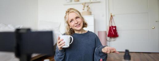 retrato de rubio joven estilo de vida bloguero, mujer registros de su hablando acerca de vida y belleza, se sienta en frente de cámara, sostiene taza, bebidas té, lo hace Blog contenido para social medios de comunicación cuenta foto