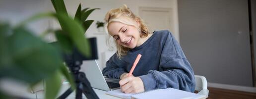 retrato de joven mujer, estilo de vida bloguero, grabación de sí misma, haciendo notas, escritura en diario, sentado en frente de ordenador portátil en un habitación y estudiando foto