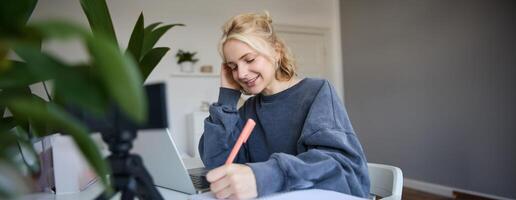 Cute smiling girl sits in a room, writes down notes, doing homework, records of herself on digital camera, creates content for vlog, lifestyle blogger doing daily routine episode photo