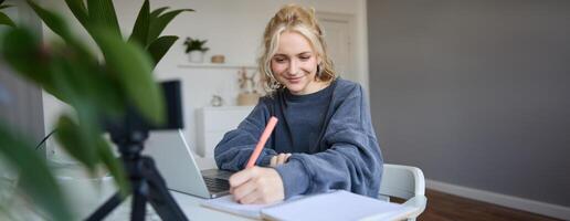 retrato de joven mujer, estilo de vida bloguero, grabación de sí misma, haciendo notas, escritura en diario, sentado en frente de ordenador portátil en un habitación y estudiando foto