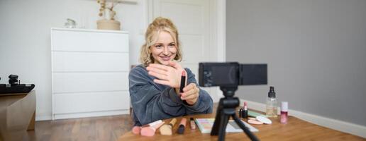 Portrait of young blond woman, teenage girl records for her social media account, shows makeup on camera, recommends lipstick to online followers, creates content in her room photo