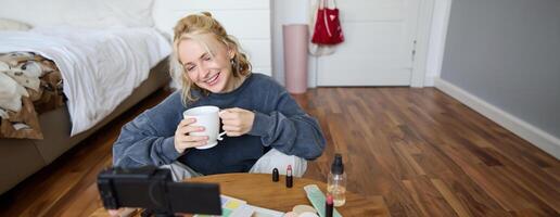 Portrait of young online influencer, social media blogger records new lifestyle in her room, has digital camera connected to stabiliser, sitting in cosy space on floor, chatting about makeup photo