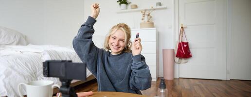 Portrait of young happy woman, content media creator recording a vlog about makeup in her room, using digital camera and stabiliser to show beauty products, holding lipstick photo