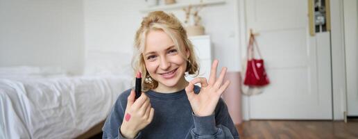 Portrait of young blond woman, content creator, recording for social media about makeup, showing okay hand sign and lipstick, recommending good cosmetic product photo