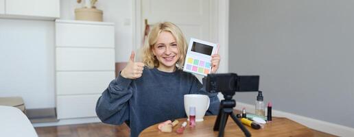 retrato de joven sonriente mujer, niña revisando paleta sombra, muestra pulgares arriba, grabación Blog, vlogging en su habitación, recomendando maquillaje producto foto