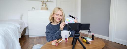 Lifestyle blogger, records in her room, has a camera on coffee table, shows eyeshadow palette to her followers, does makeup tutorial, vlogger working indoors, creates content for social media photo