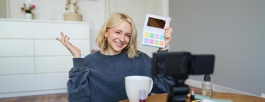 Lifestyle blogger, records in her room, has a camera on coffee table, shows eyeshadow palette to her followers, does makeup tutorial, vlogger working indoors, creates content for social media photo