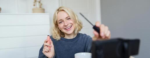 Image of cheerful, beautiful young lifestyle blogger, woman sitting on floor and recording about makeup, holding mascara, making lifestyle content for her social media account and followers photo