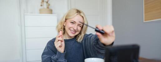 Portrait of young smiling woman in her room, recording on camera, lifestyle vlog for social media, holding mascara, reviewing her makeup beauty products, showing how to use cosmetics photo
