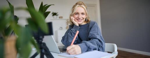 Cute smiling girl sits in a room, writes down notes, doing homework, records of herself on digital camera, creates content for vlog, lifestyle blogger doing daily routine episode photo