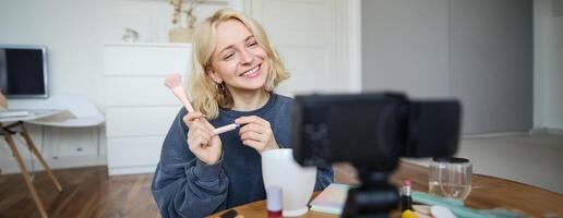 Smiling young beautiful lifestyle blogger, girl records a beauty vlog in her room, using professional camera, showing makeup cosmetics to her followers, creates content for social media photo