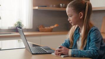 in movimento telecamera caucasico ragazzo ragazza scolara ascolta in linea lezione scuola classe a casa scrittura compiti a casa. quarantena lontano formazione scolastica. bambino figlia studiando a distanza con il computer portatile e-learning a cucina video