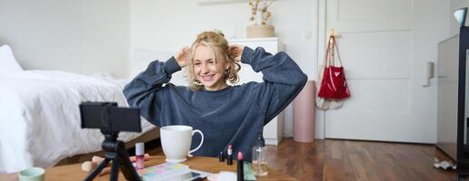 retrato de linda rubio chica, estilo de vida blogger se sienta en piso con cámara y estabilizador, muestra cómo a hacer peinado y constituir, lo hace casual vlogging para social medios de comunicación, se sienta en habitación foto