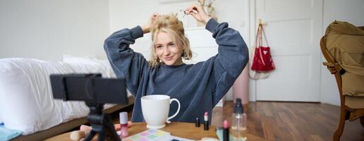 Lifestyle blogger records of herself while doing hairstyle, showing makeup lifehacks for social media followers, vlogging, using camera for her blog, sitting in a room, drinking tea photo