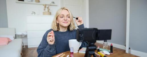 Image of cheerful, beautiful young lifestyle blogger, woman sitting on floor and recording about makeup, holding mascara, making lifestyle content for her social media account and followers photo