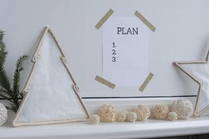 PLAN checklist text on paper note stick on while wall with cozy minimalistic handmade Christmas decor. New year aims resolutions. Low key festive Planning and setting goals concept photo