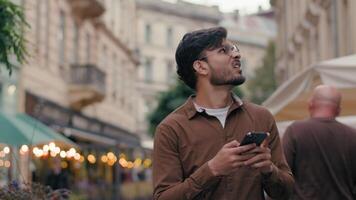 unzufrieden indisch arabisch ethnisch männlich Reisender Mann Kerl Tourist erkunden Stadt Route Reisen draußen. Geschäftsmann Bummel im Ausland Straße suchen Handy, Mobiltelefon Telefon Gadget Handy gehen Stadt, Dorf mit online Karte video