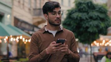 Young Indian Arabian ethnic man male traveler tourist guy businessman walking city street holding mobile phone using modern technology online travelling service chatting message looking around outside video