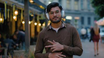 Indian Arabian male man teen gen z happy guy student businessman tourist in modern wireless headphones having fun listen relax music song moving walk gesticulate dance outdoors night city town street video