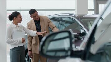 africain américain femme vendeur Marchand Commerçant consultant dans auto salle d'exposition Aidez-moi homme client choisir Nouveau voiture moderne électrique voiture vendeuse consulter Explique véhicule Caractéristiques vente dans concession boutique video