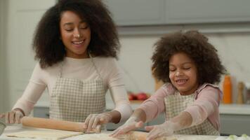 alegre africano americano madre mujer enseñar pequeño hija niño niño niña hacer aplastamiento masa con rodillos en cocina contento familia utilizar ecológico productos ingredientes Cocinando juntos sonriente video