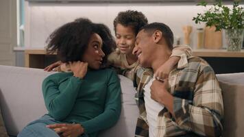 Happy joyful African American family adult parents married couple mother father little boy adorable child kid son having fun talking cuddle hug childcare togetherness indoor home cozy modern apartment video