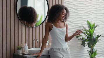 africano americano feliz sorridente mulher usando Móvel telefone navegando conversando Smartphone menina Telefone social meios de comunicação SMS mensagem compras conectados Internet surfar aplicativo dentro banheiro casa beleza procedimento video