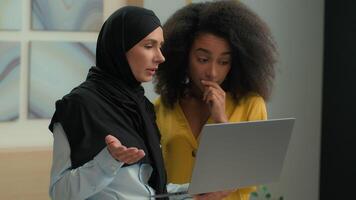 Two diverse female colleagues business partners coworkers multiracial African American muslim in hijab women girls businesswomen talk discuss online project laptop working together in corporate office video