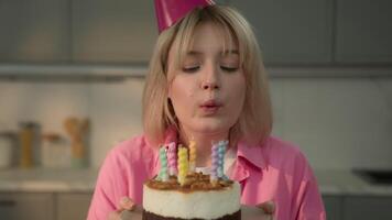 Caucasian girl smiling joyful happy excited young woman lady alone female smile carefree in festive cap hat holding cake blowing candles wish holiday birthday celebrating at home apartment insurance video