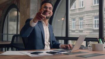 Indian business man manager CEO employer working laptop at corporate office workplace happy joyful Arabian male businessman pointing finger at camera hey you hand gesture positive cheerful guy smile video