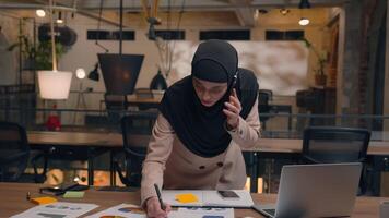 Muslim woman multitasking talk mobile phone call discuss business project female Arabian islamic girl hijab businesswoman designer organizer planning manage tasks write notes on table office workplace video