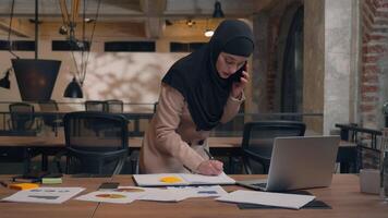 Muslim islamic Arabian woman girl in hijab working laptop project analysis planning female smile businesswoman talking mobile phone stand at table write notes speak smartphone business call in office video