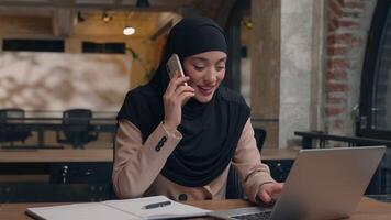 Muslim Arabian islamic woman in hijab happy businesswoman talk mobile phone work at corporate company office female consultant manager business girl consult client online using laptop write task smile video