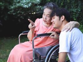 Happy Asian senior woman sitting on wheelchair with her son in the garden. talking and pointing. photo