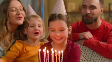 Caucasian family sing happy birthday little girl child daughter blow cake candles wish celebration funny joyful parents kids children siblings sisters in festive hats celebrate birth event party home video